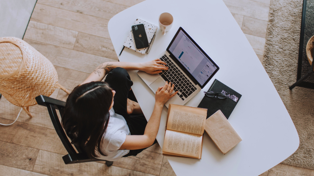 A person sits at a white desk working on a laptop with the Buffer for Business tutorial on the screen, surrounded by open books, a notebook, a smartphone, a cup of coffee, and glasses, suggesting a focused learning environment.