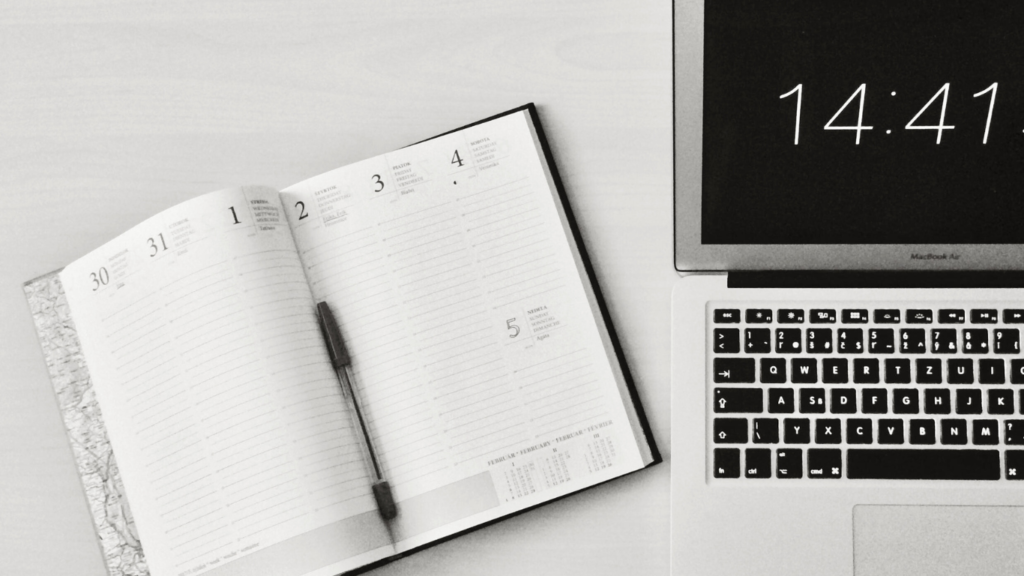 Black and white image of an open planner with a pen on the left and a laptop displaying the time 14:41 on the right, symbolizing time management and scheduling in digital marketing.