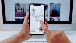 Close-up of a person's hands holding a smartphone with the screen displaying a social media profile as part of a Buffer for Business Tutorial. The phone is positioned against a computer monitor, illustrating the process of managing and coordinating social media content across devices for streamlined digital marketing.