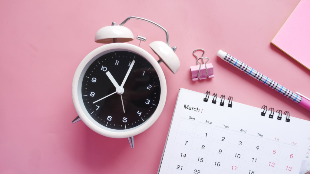 An overhead view of a white alarm clock, pink binder clips, a pencil, and a calendar on a pink background.