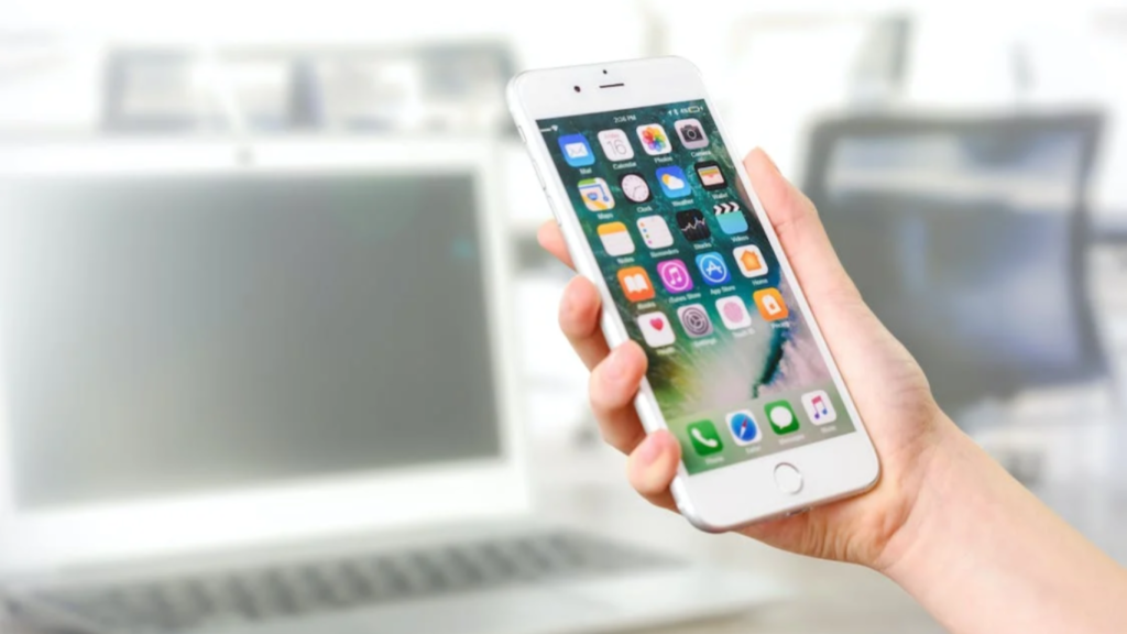 A person's hand holding an iPhone with a display full of various colorful app icons, blurred office background with a laptop and chair, symbolizing modern mobile technology use in everyday life.