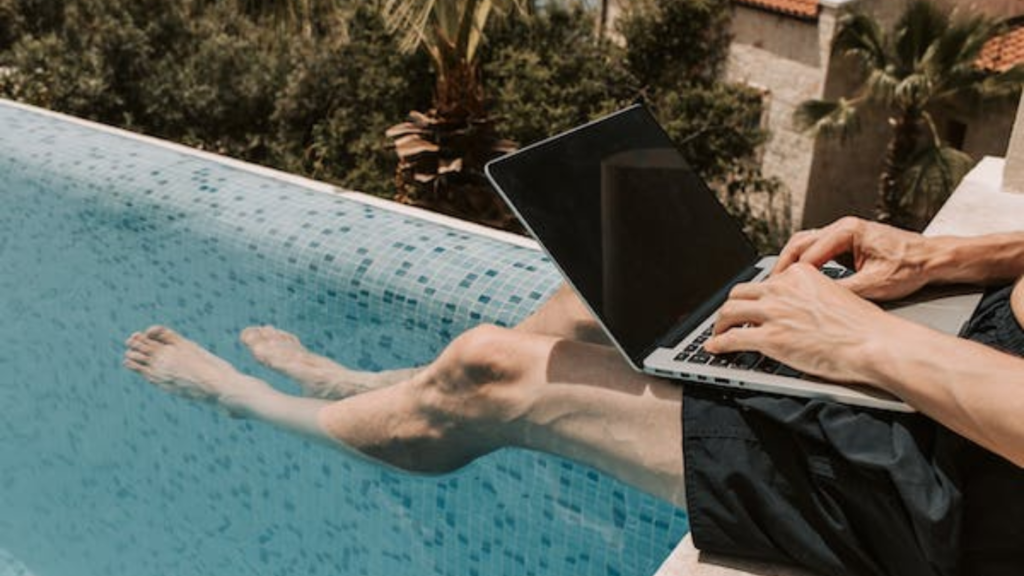 A person casually working on a laptop with their feet dipped in a swimming pool, symbolizing the relaxed lifestyle of a travel blogger creating engaging content.