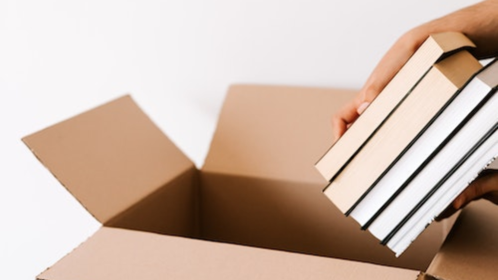 An open cardboard box with a hand placing a stack of neatly aligned books inside, symbolizing the potential to 'Make Money Reading Books' by offering e-book formatting and design services to authors and publishers seeking to digitalize their manuscripts professionally.