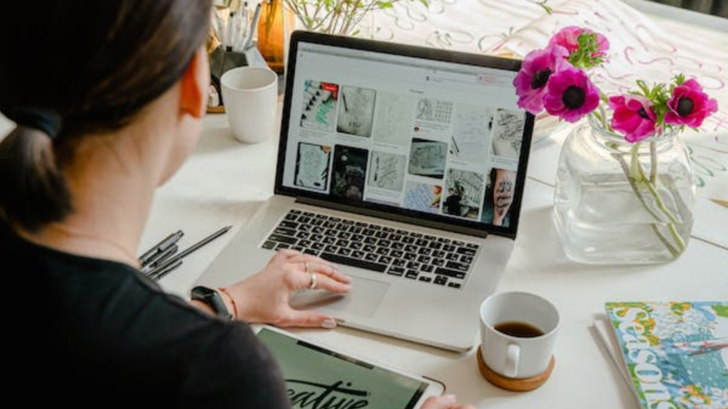 A person from behind working on a laptop with a webpage displayed, surrounded by creative tools including a notebook, a pen, a cup of coffee, and a vase with pink flowers, indicating a productive content updating session.