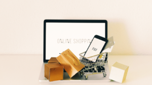 A small shopping cart filled with boxes and a smartphone displaying 'PAY' on its screen, against the backdrop of a laptop with 'ONLINE SHOPPING' on the display, symbolizing online shopping and payment, for a guide on Holiday Shopping Savings Apps.