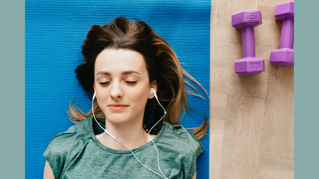 A woman is relaxing on a blue yoga mat with her eyes closed, listening to music through white earbuds. Next to her are two purple dumbbells on a wooden floor. This scene represents how artists and musicians can listen to music and get paid online by having their tracks streamed on platforms like Spotify, Apple Music, and Amazon Music.