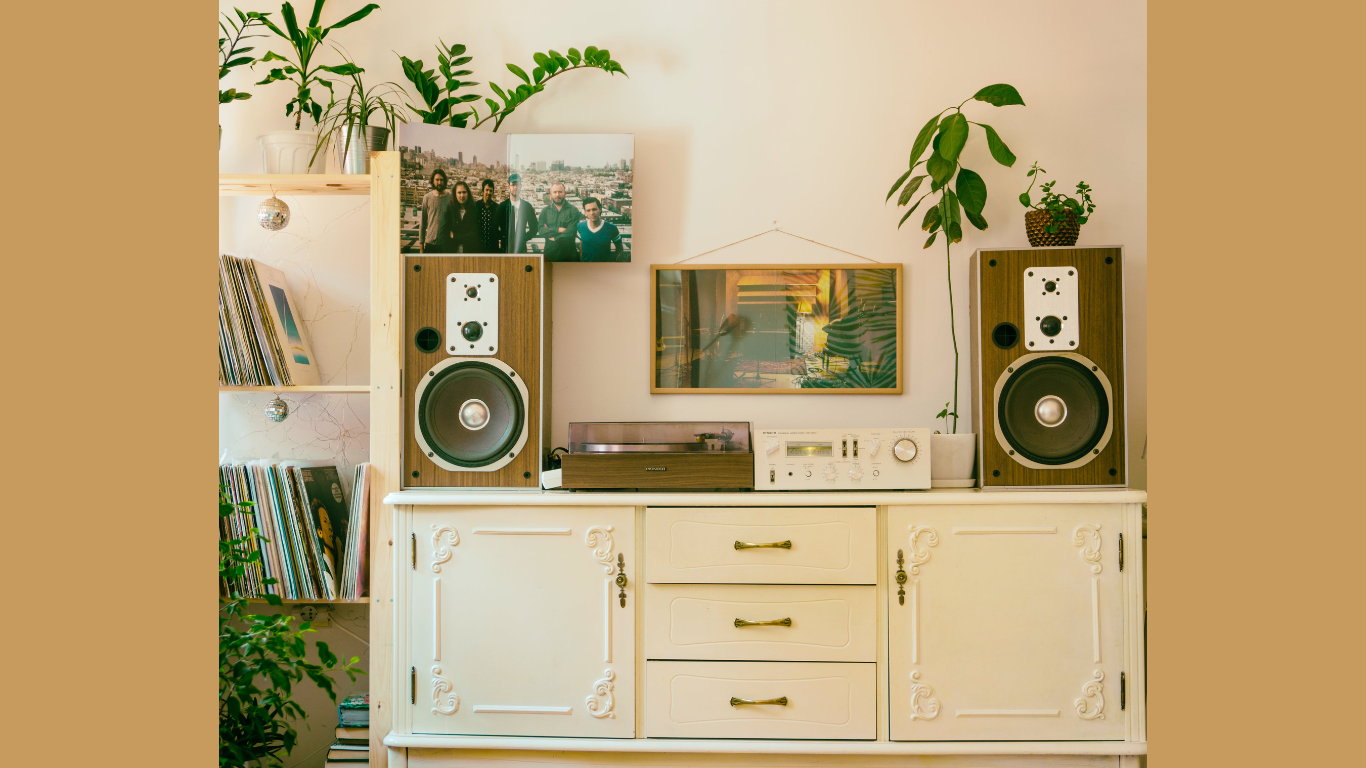 A cozy and vintage music listening setup in a living room, featuring a classic white dresser with an old-school silver turntable on top, flanked by two large wooden speakers. Above the turntable hangs a framed picture of a record player, and on the dresser’s shelves rests a collection of vinyl records beside potted plants. This serene space embodies the pleasure of music listening that can also be a source of income when paired with online paid music platforms.