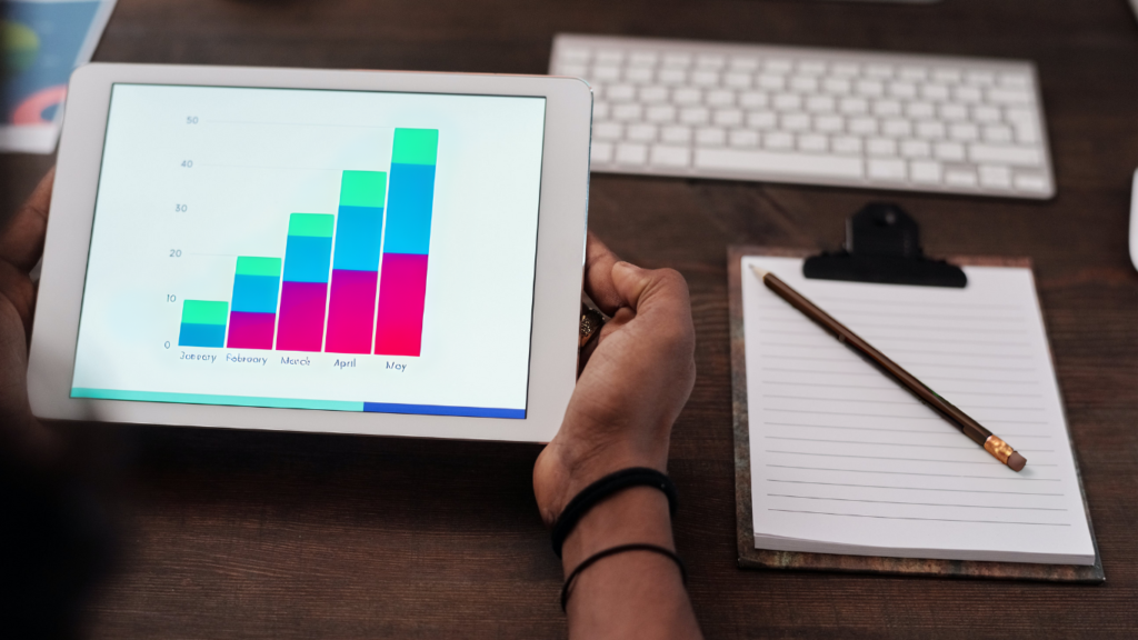 Hands hold a tablet showcasing a bar chart with month-over-month growth, reflecting data analysis for 'How to Improve Website SEO'. On the desk, a notepad and pencil suggest strategic planning, alongside a keyboard, ready for implementing SEO improvements.