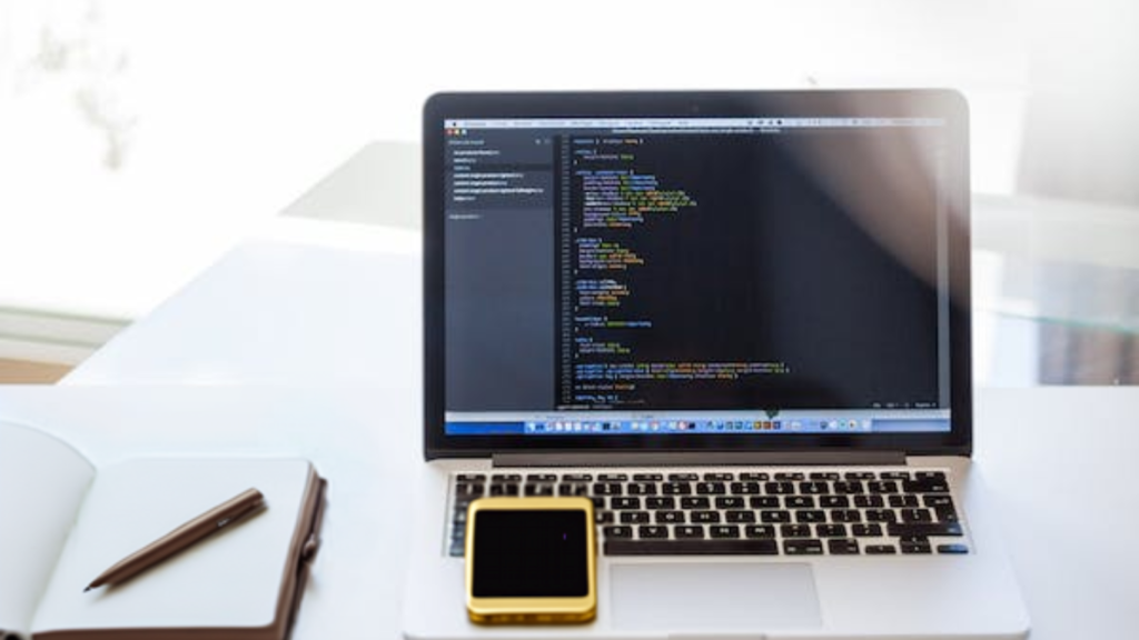 Laptop on a white desk displaying HTML code, indicative of 'How to Improve Website SEO', with a notebook and pen to the side for strategy notes, and a smartphone, suggesting a multi-device approach to optimizing web content.