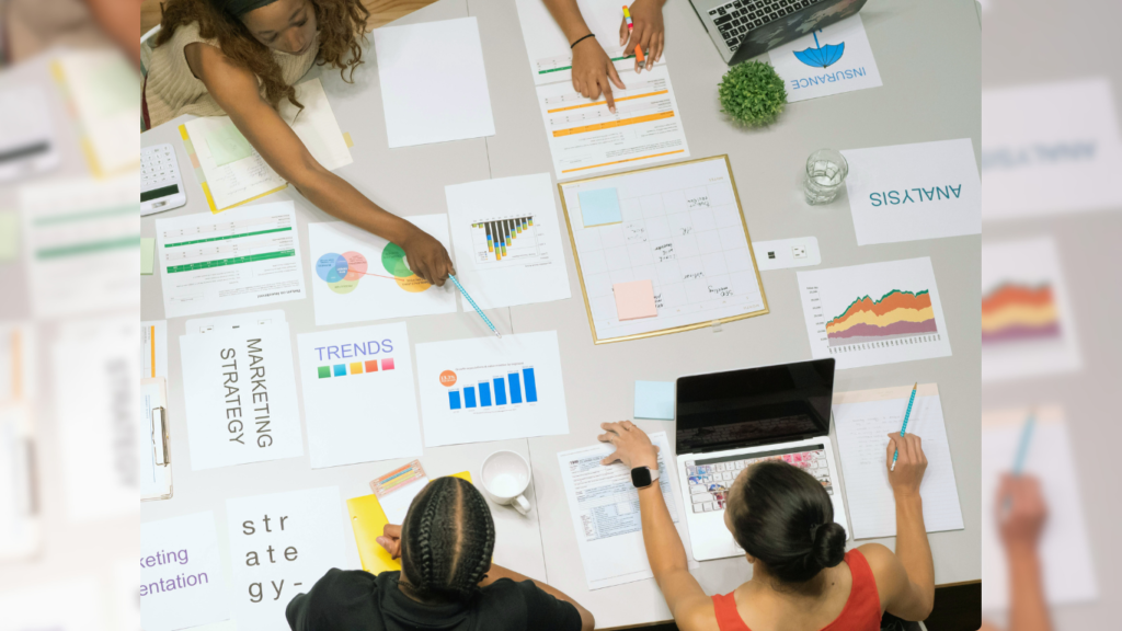 A top-down view of a diverse group of professionals working around a table covered with marketing strategy documents, graphs, and a laptop, actively engaged in a collaborative SEO planning session.